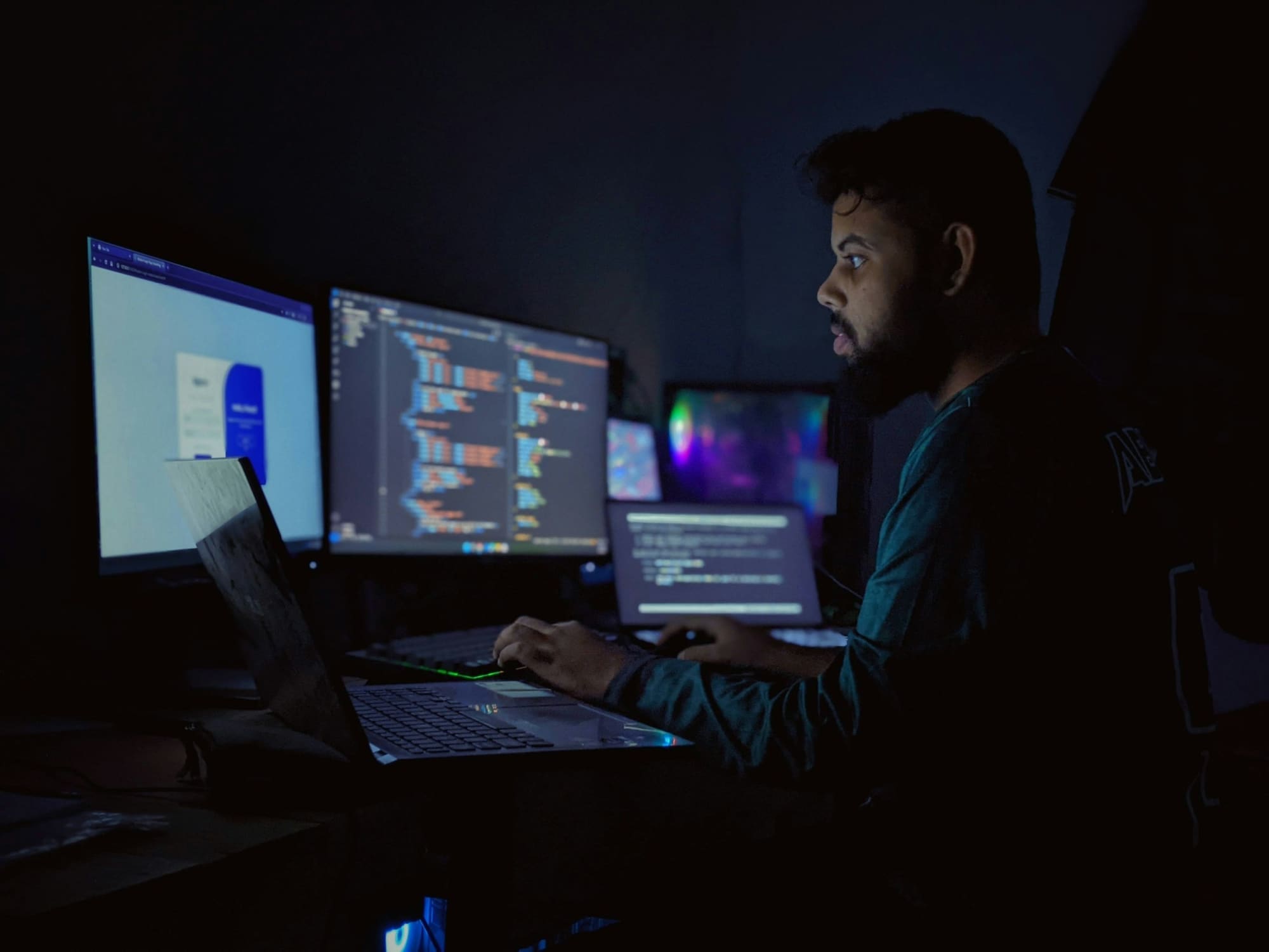 A developer works late in a dark room in front of several monitors showing code.