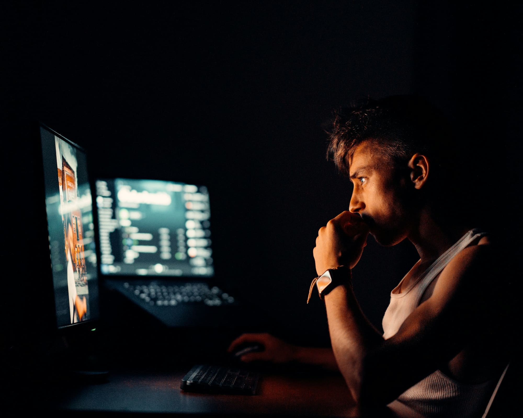 A creator sits in a dark room, face lit by two monitors, chin resting on one hand while studying the screen.