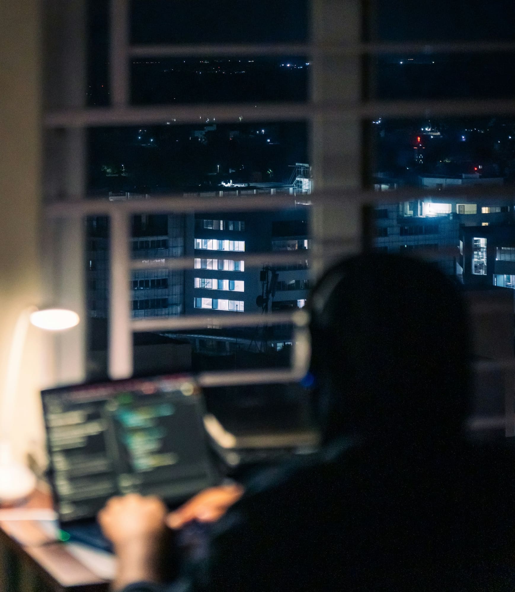 A person works on a laptop at night beside a window overlooking city lights.