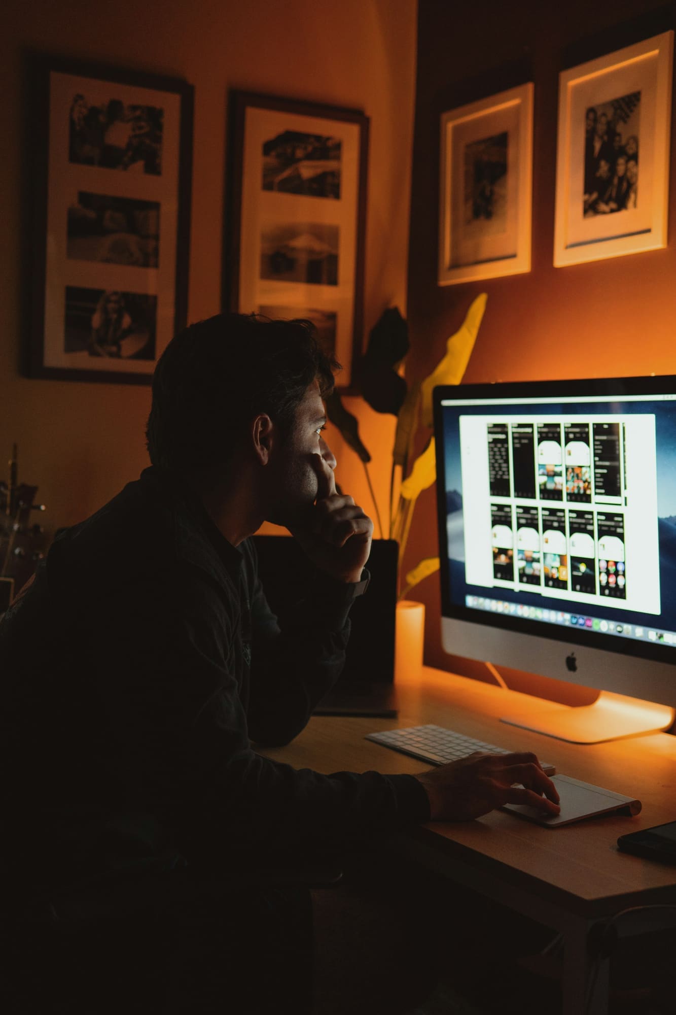 A photographer at a desk in a warmly lit room reviews a grid of photos on a desktop monitor, with framed prints on the wall behind.