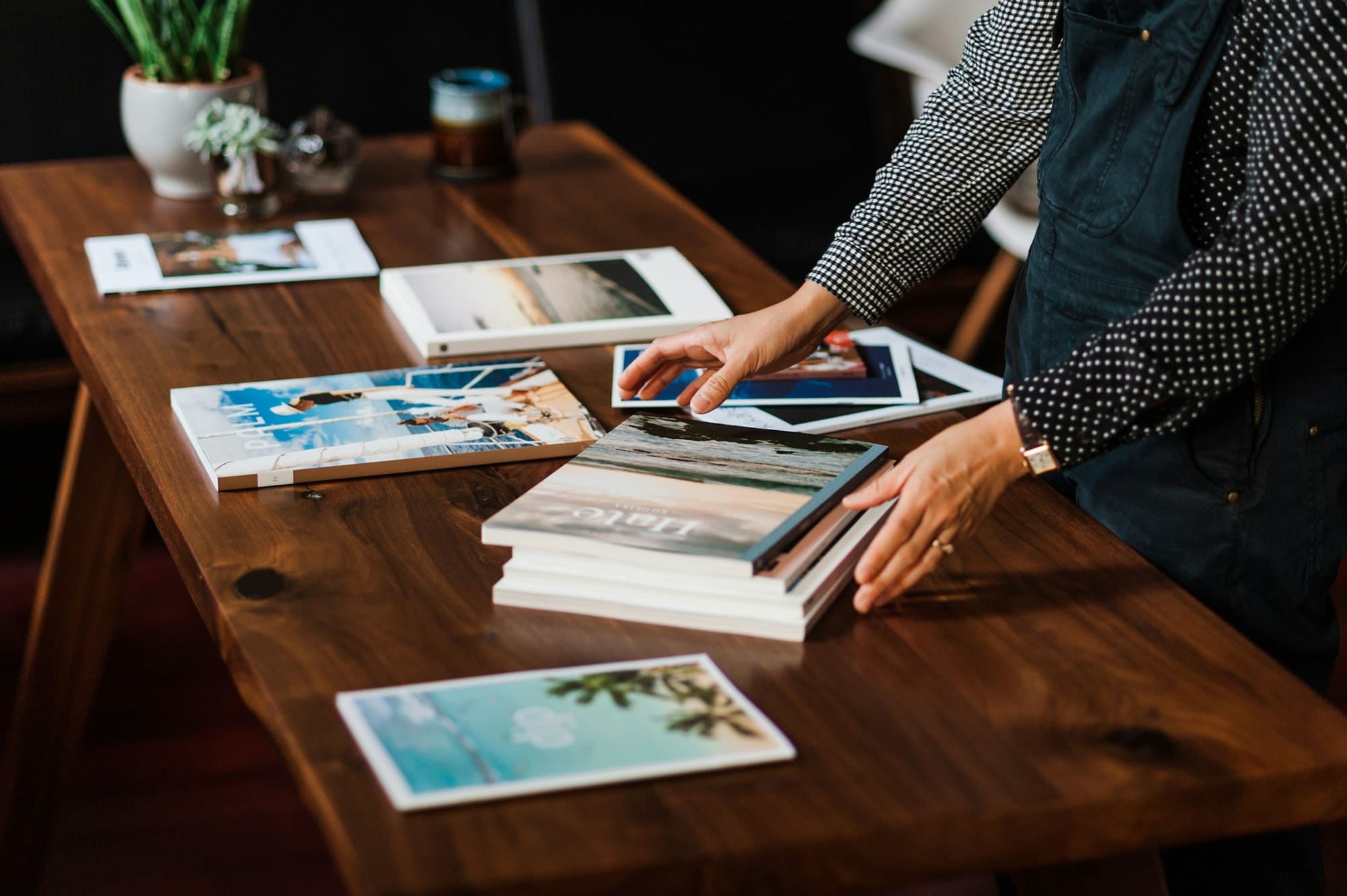 A person arranges printed photo books and prints on a walnut table.